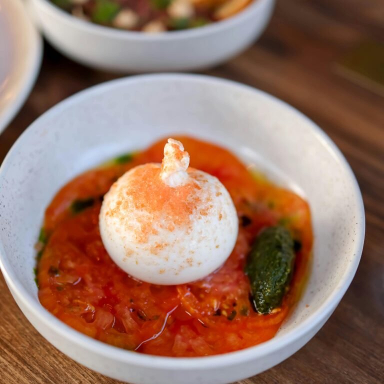Close-up of burrata cheese atop sliced tomatoes in a white bowl on a wooden table.